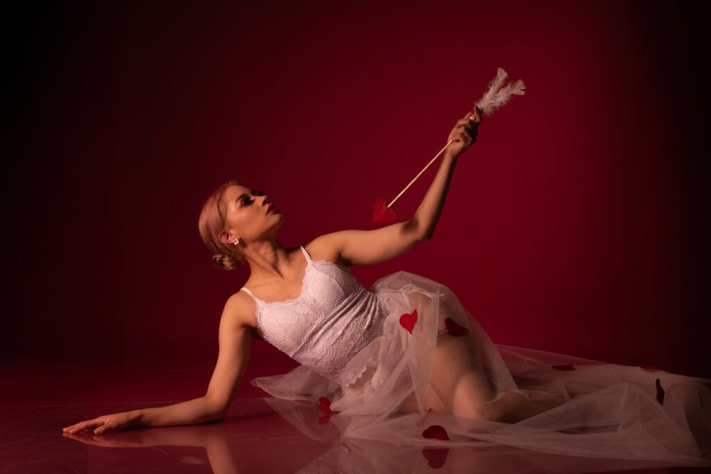 A graceful dancer posing with a cupid-themed arrow against a dramatic red backdrop.